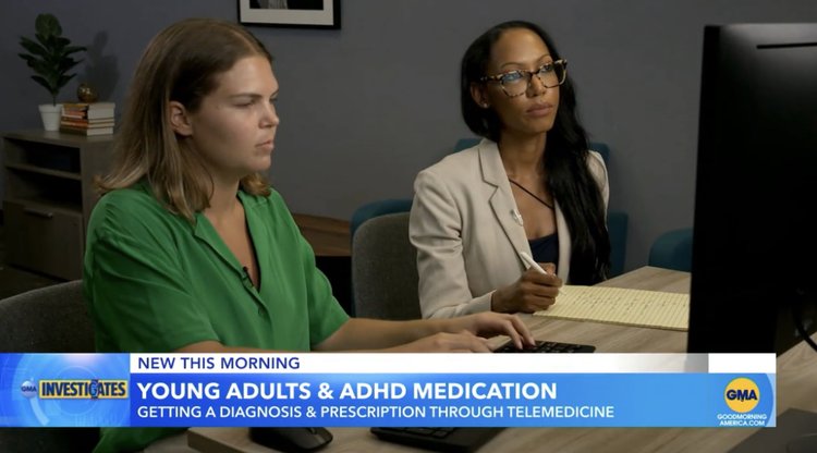 Two women, one in a green blazer, the other in a gray blazer, sit at a desk looking at a computer screen with a news banner about adhd medication and telemedicine.