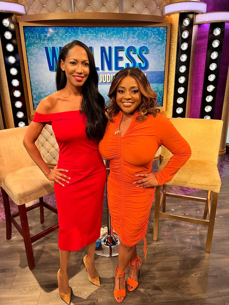 Two women stand smiling in front of a tv show set titled witness with a lavish purple background one is in a red dress, the other in an orange dress, both elegant