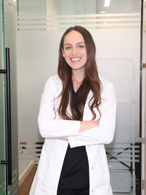 A smiling woman with long brown hair, wearing a white lab coat and black shirt, stands confidently with her arms crossed in an office setting with glass panels.