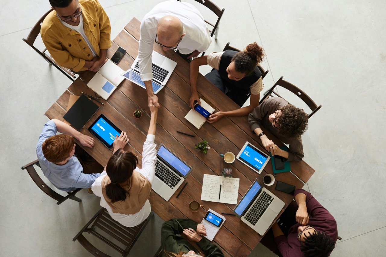 Top view of a diverse group of professionals engaged in a meeting around a rectangular table, using laptops and discussing documents.
