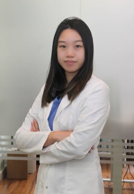 A young asian female medical professional stands confidently with arms crossed, wearing a white lab coat over a blue blouse in a clinical setting.