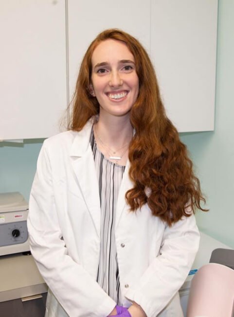 A young female scientist with long curly red hair, wearing a white lab coat and gloves, smiling in a laboratory setting.