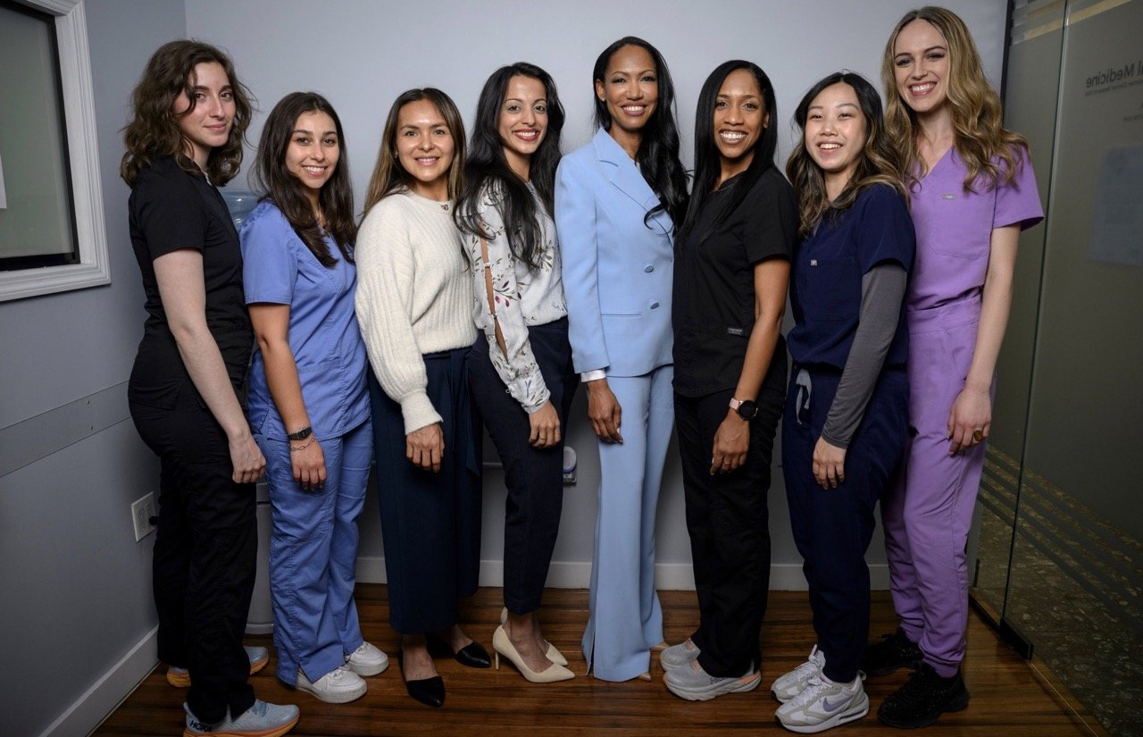 Group of seven women, presumably healthcare professionals, smiling and standing together in a hallway. they are wearing a mix of scrubs and casual business attire.
