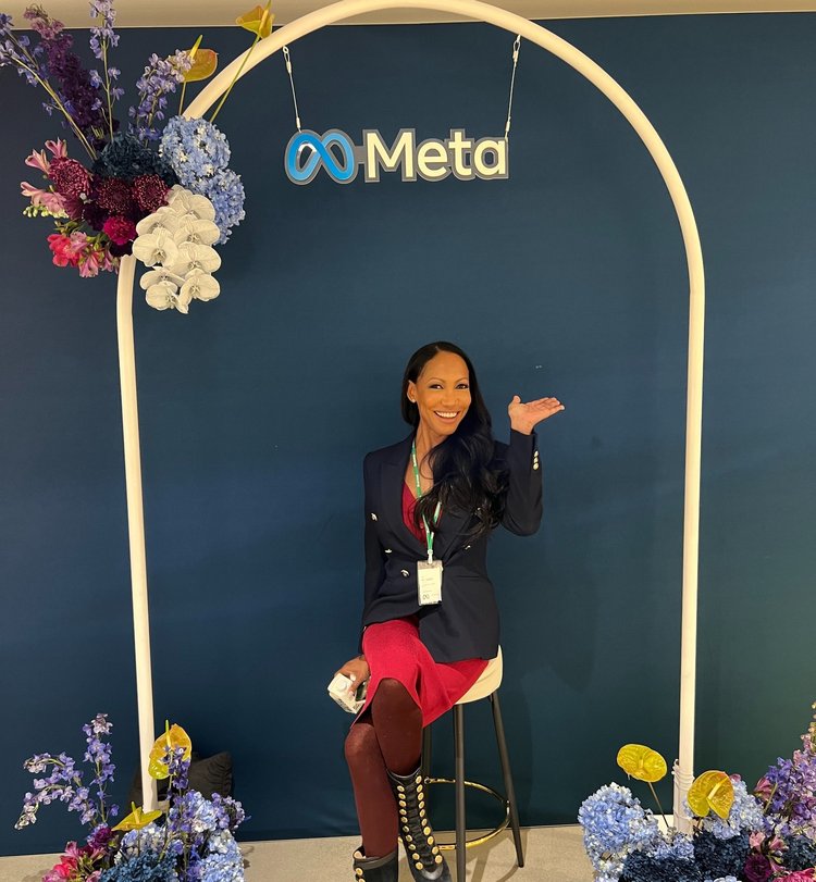 A woman in a navy blazer and red skirt is smiling and posing with one hand raised, sitting under an arch with the meta logo and decorated with colorful flowers.