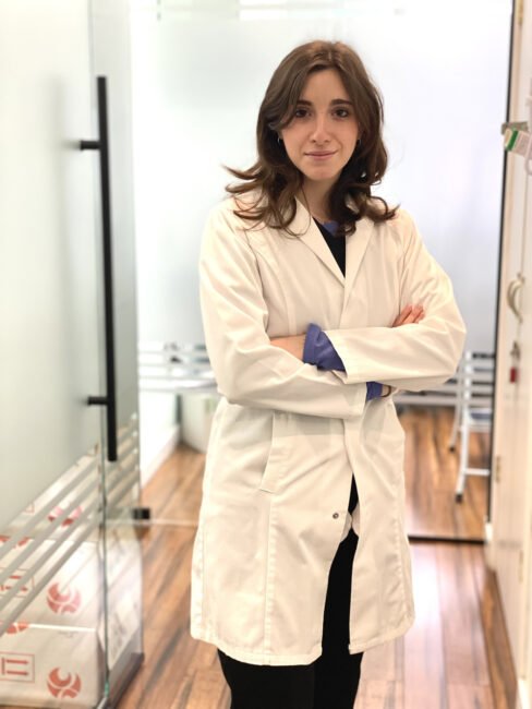 A young female scientist with brown hair, wearing a white lab coat and latex gloves, stands in a laboratory, arms crossed, smiling at the camera.