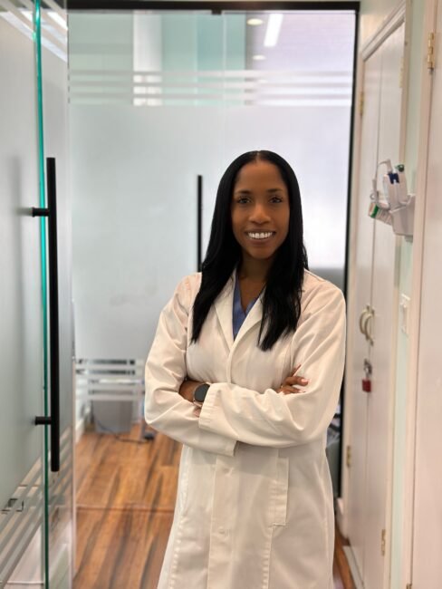 A confident woman in a white lab coat stands with crossed arms, smiling in a clinical setting with glass doors and medical equipment in the background.