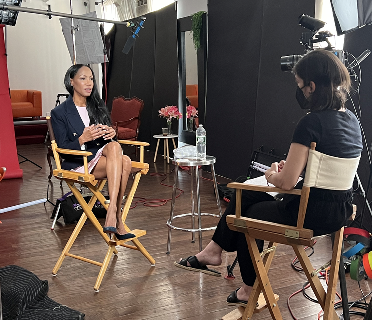 Two women sit in director's chairs in a studio setting, engaged in an interview. the interviewee, dressed in black, speaks while the interviewer takes notes. cameras and lighting equipment surround them.