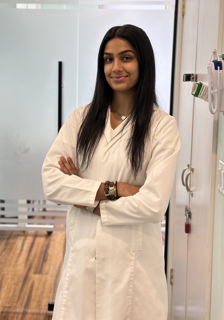 A confident young female scientist wearing a lab coat stands with crossed arms in a laboratory setting, smiling slightly at the camera.
