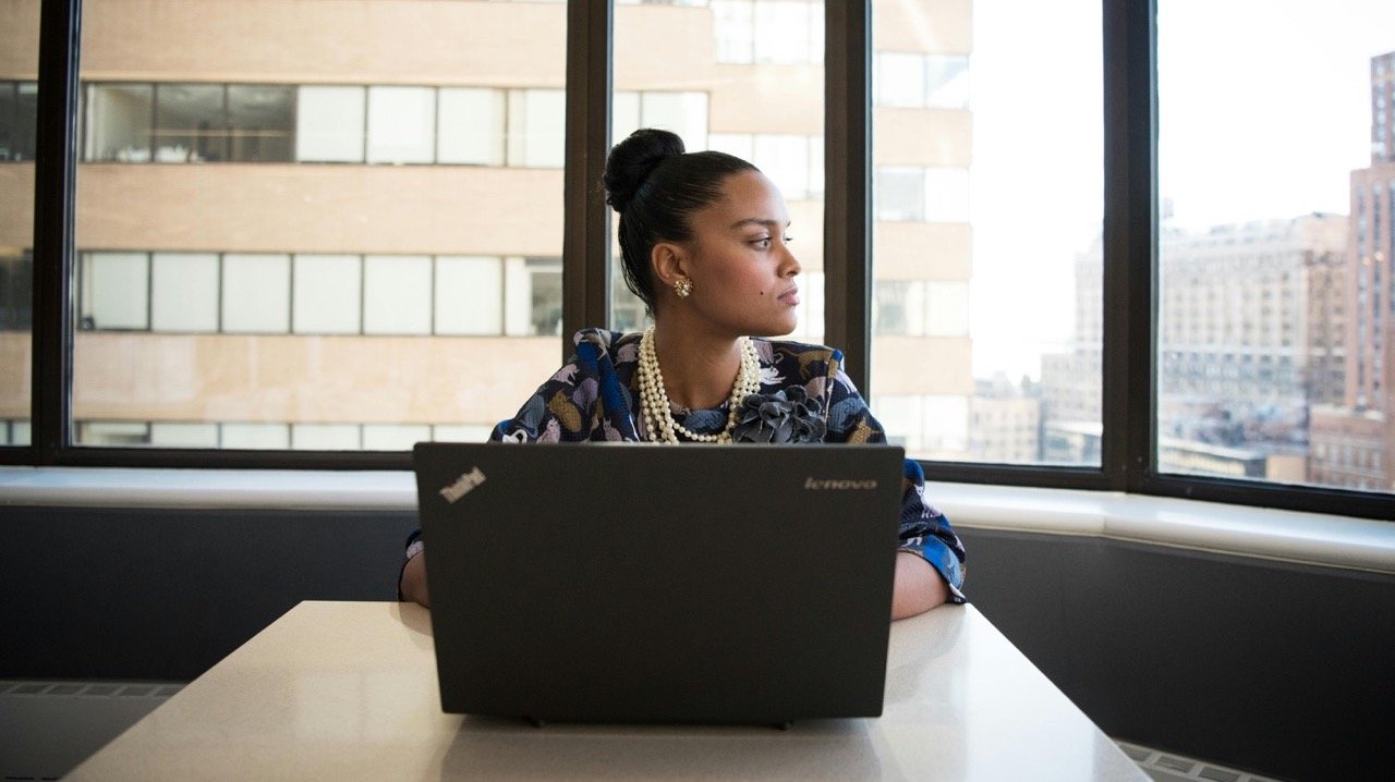 A woman sitting at a desk with a laptop, looking out a large window towards a cityscape, reflective and thoughtful.